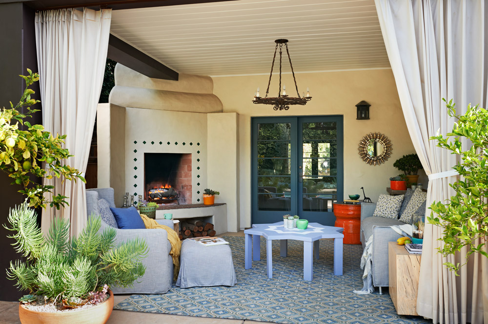 The drapery and potted plants frame an outdoor living room in blues and greys. A patterned fiber rug defines the area which features a welcoming fireplace, while the lounge chair and ottoman hug the corner near one of the posts.